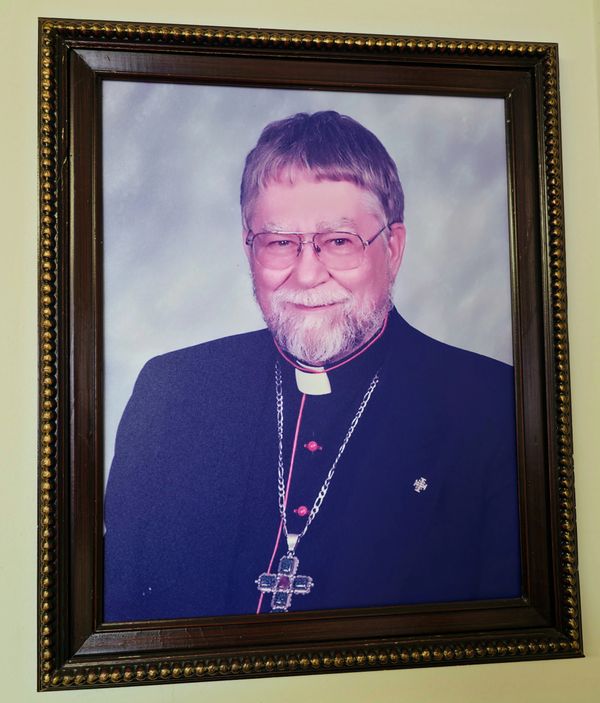 Framed portrait of a smiling clergyman wearing glasses and religious attire with a large cross necklace.