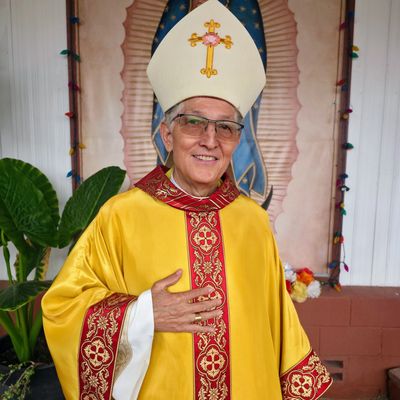 A smiling bishop in yellow and red religious robes and mitre stands indoors.