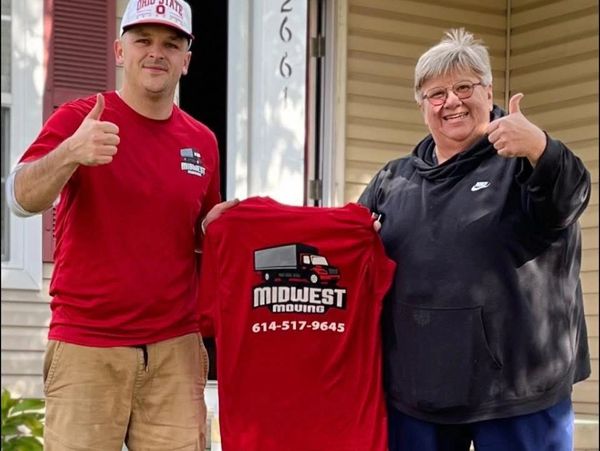 Two people smiling and giving thumbs up in front of a house, holding a red Midwest Moving shirt.