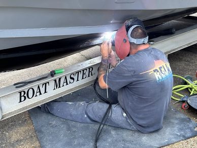 A man welds the underside of a boat on a trailer labeled 'BOAT MASTER'.