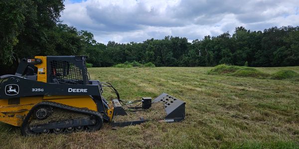 A John Deere 325G compact track loader clearing a grassy field under a cloudy sky.