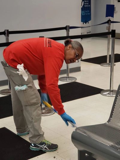 A woman in a red shirt and blue gloves cleaning the floor next to a metal bench