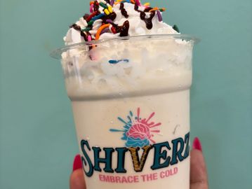 Hand holding a creamy milkshake topped with whipped cream, chocolate drizzle, and colorful sprinkles.