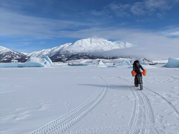 Person biking on snowy terrain with mountains and icebergs in the background.