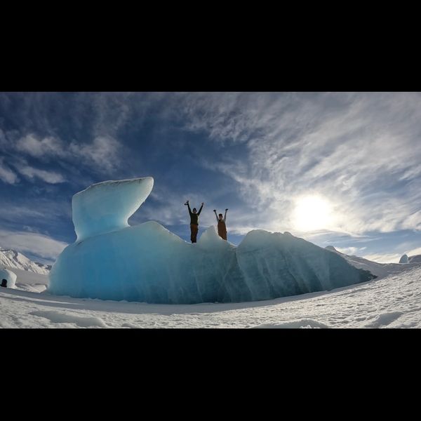 Two people celebrating on a large blue ice formation under a bright sky.