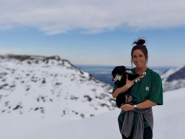 A woman holding a small dog in snowy mountains with a smile.