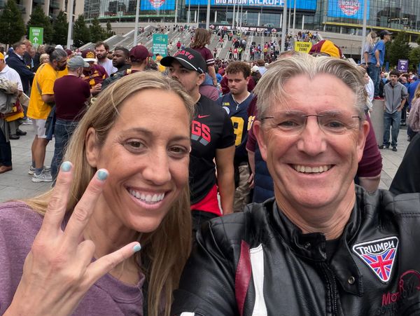 Two people smiling and posing for a selfie in a crowded outdoor event.