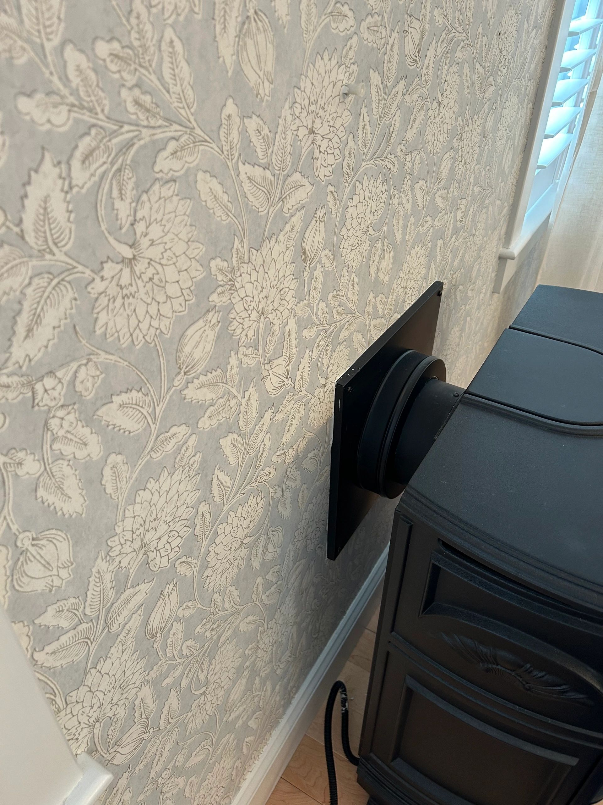 Black stove pipe venting through floral wallpapered wall.