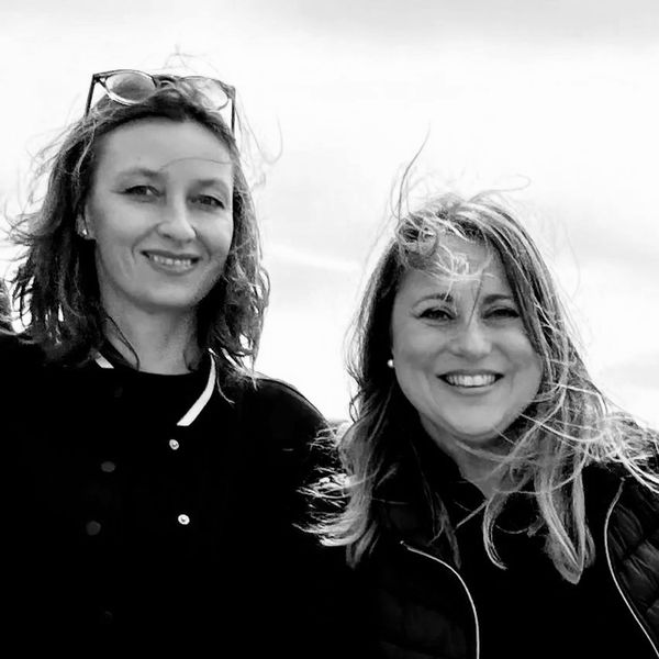 Two smiling women standing close together outdoors, wind blowing their hair.
