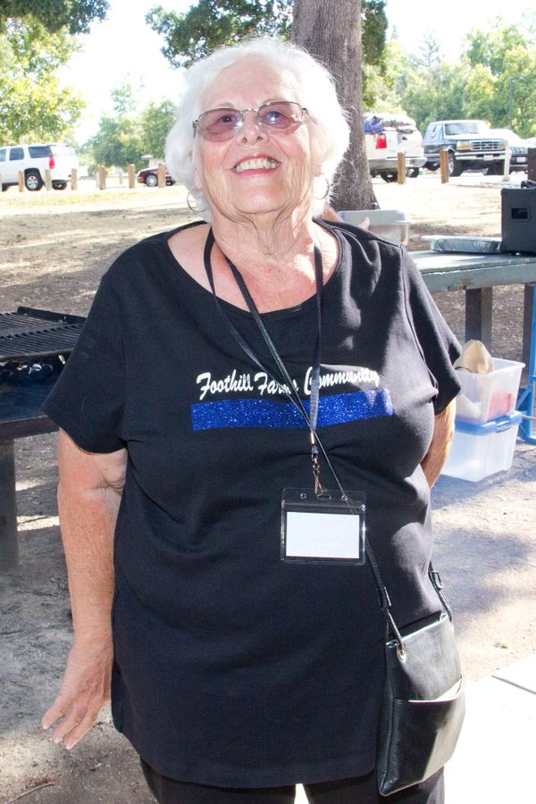 Elderly woman smiling outdoors wearing a black Foothill Farms Community shirt and name badge.