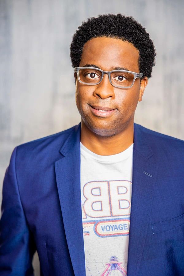 Man with glasses wearing a blue blazer and printed white t-shirt, smiling.