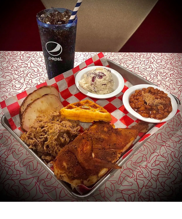A barbecue platter with chicken, pulled pork, sliced turkey, waffle, baked beans, potato salad, and a Pepsi.