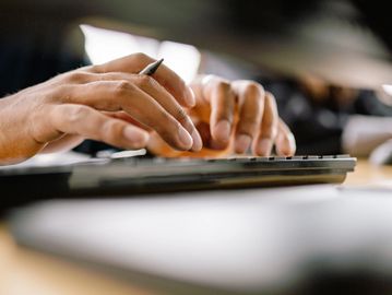 Close-up of hands typing on a keyboard with a pen in one hand.