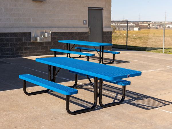 Blue outdoor picnic tables on a concrete patio near a building and water fountains.