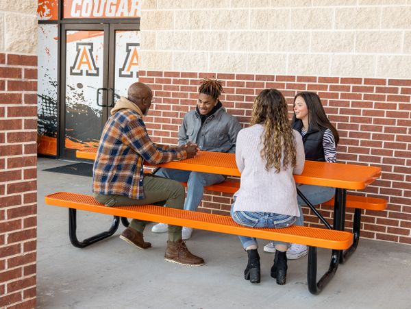 Four people sitting and chatting at an orange picnic table outside a building.