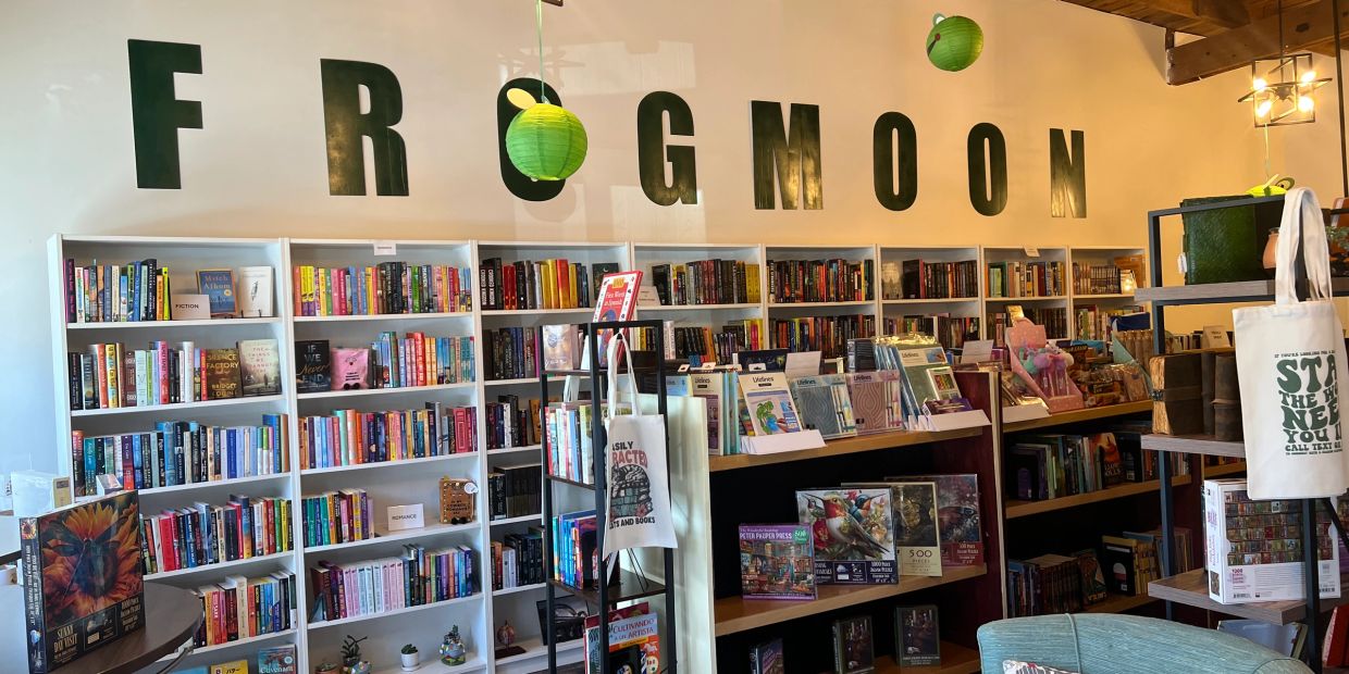 Cozy bookstore named FROGMOON with shelves full of books and decorative green lanterns.