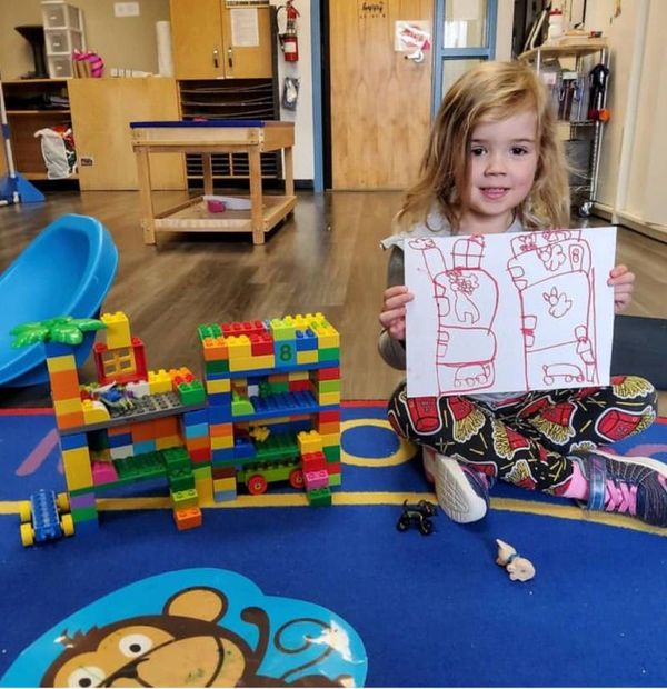 A child holding up their drawing with their Duplo Lego creation on the floor beside them. 