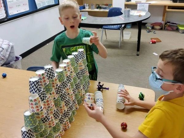 Two children standing beside a table, building a  wall out of small paper cups on the table top. 
