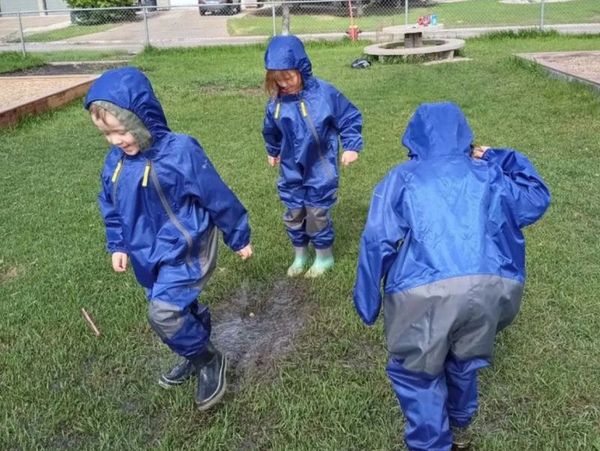 A small group of children in rain suits and rubber boots, jumping in puddles.  