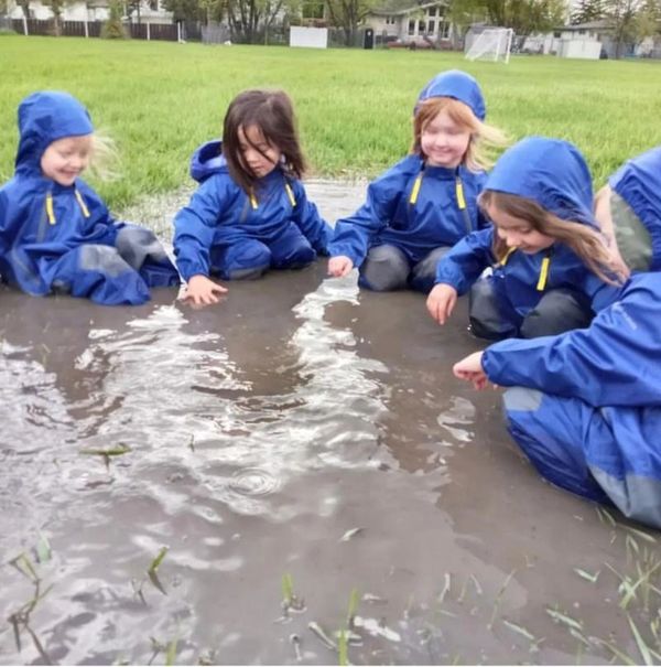 A small group of children in rain suits kneeling in a large puddle.