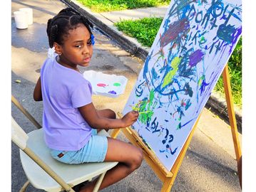 Young girl painting on an easel outdoors in bright daylight.