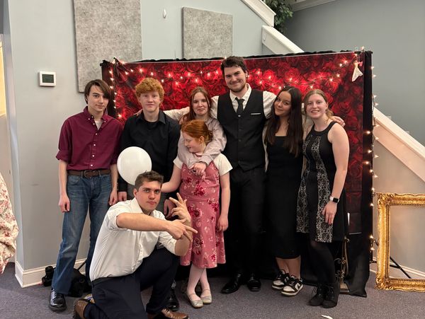 A group of seven young people posing in front of a red rose backdrop with string lights.