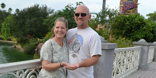 A couple smiles while standing on a bridge near water and greenery.