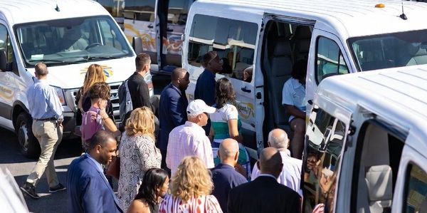People boarding white and blue shuttle vans in a parking area on a sunny day.