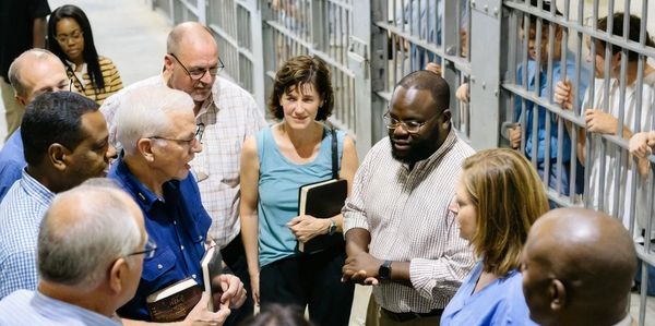 Group of people having a discussion in a prison hallway with inmates behind bars.