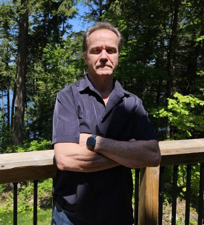 Man standing with arms crossed on a wooden deck surrounded by trees.