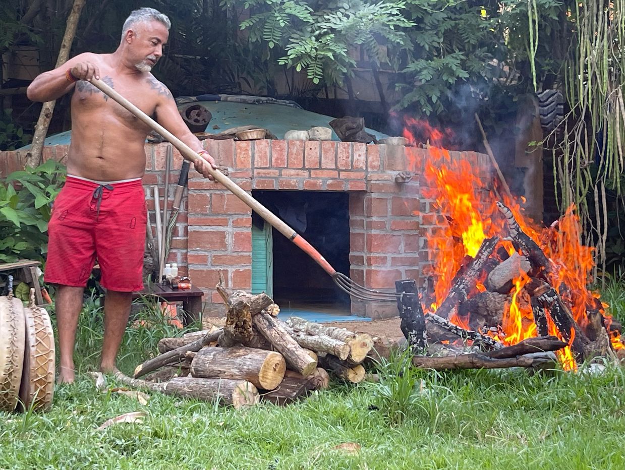 Man tending a bright outdoor fire with a long wooden tool near a brick structure.