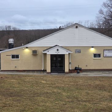 A simple beige community building with a white roof and black double doors under a cloudy sky.