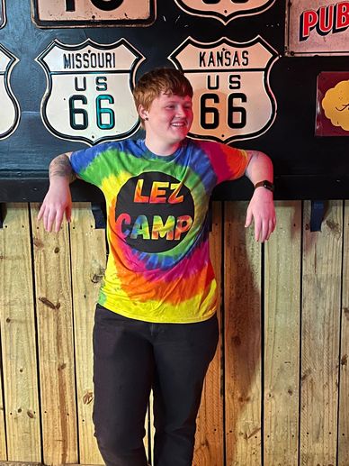 Person in a colorful tie-dye "LEZ CAMP" shirt standing by wooden wall with Route 66 signs.