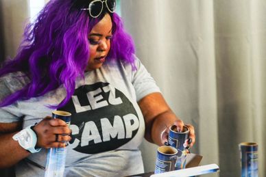 Person with purple hair reading and holding cans at a table indoors.