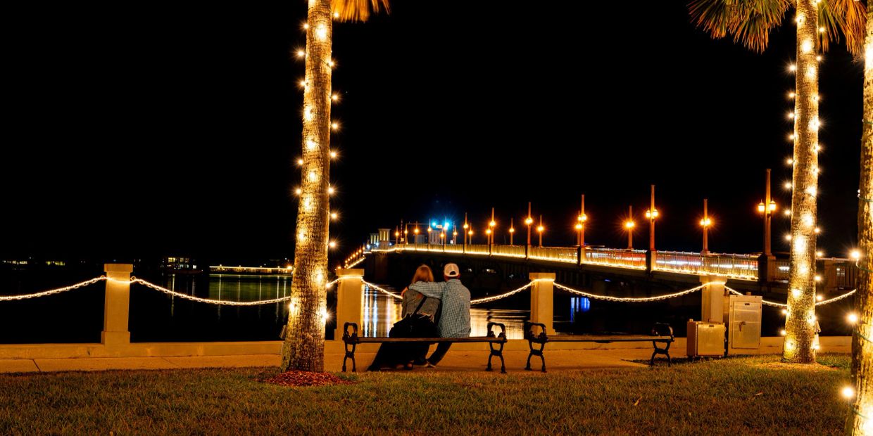 Couple sitting on bench by a lit bridge at night.