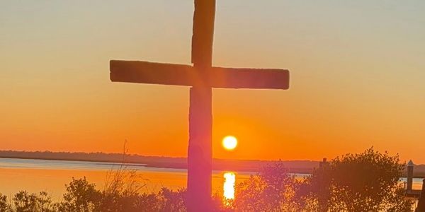 Silhouette of a cross against a vibrant sunset over water.