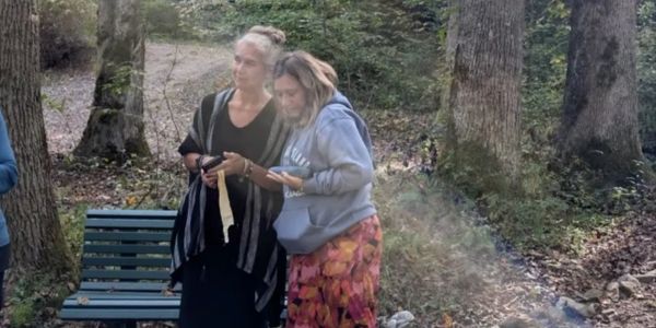 Two women outdoors near a bench in a wooded area, looking at a phone.