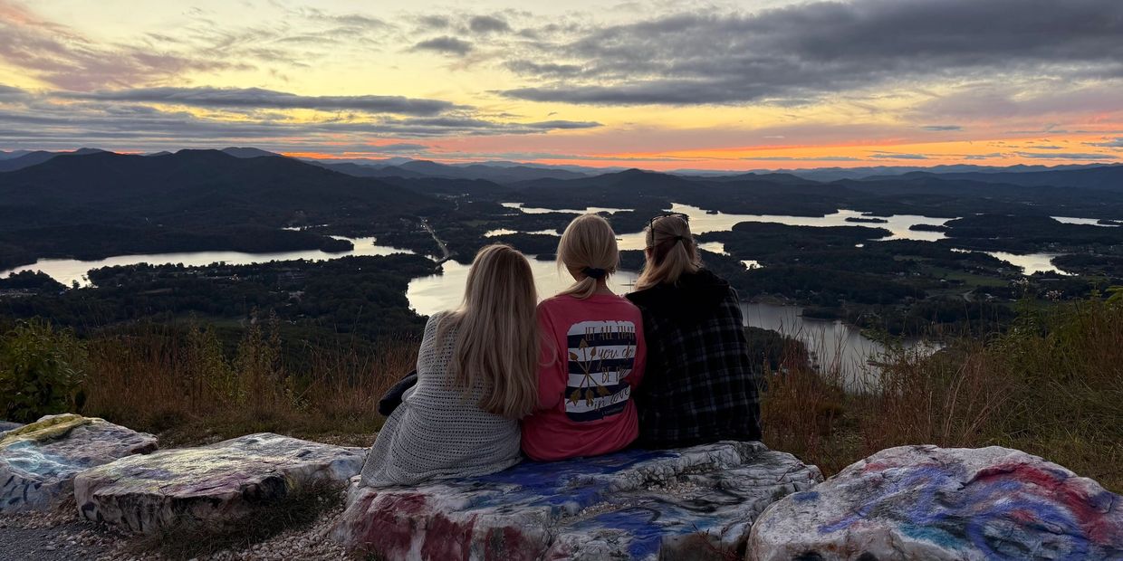 Three friends sitting on a rock overlooking a scenic sunset over lakes and mountains.