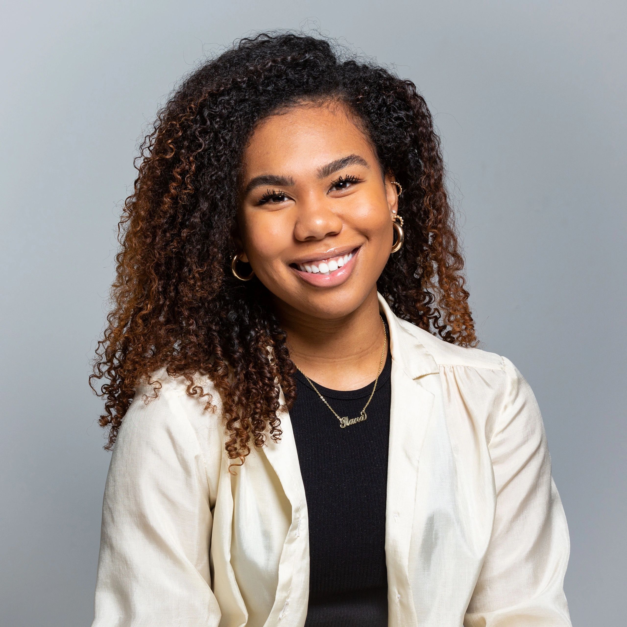 Smiling woman with curly hair wearing gold hoop earrings and layered necklaces.