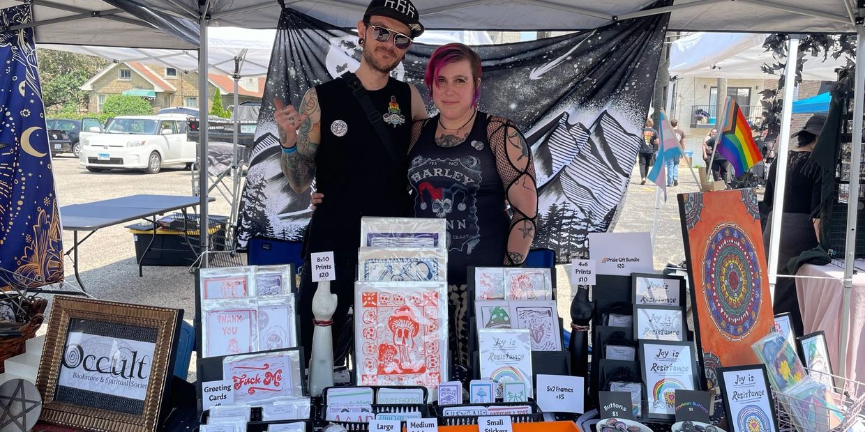 Two people stand behind a booth selling occult-themed prints, cards, and buttons at an outdoor market.