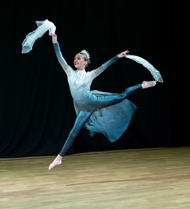 Ballet dancer leaps gracefully on stage with flowing costume and scarf.