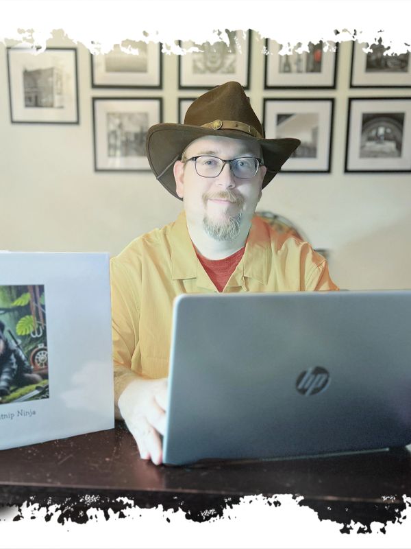 Man in a brown hat working on a laptop at a desk with framed photos behind.