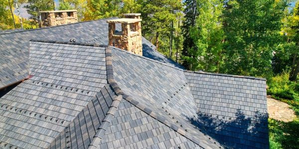 A complex roof with grey shingles and stone chimneys surrounded by trees.