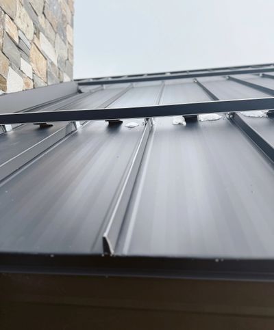 Close-up of a gray metal roof with a stone wall beside it under a cloudy sky.