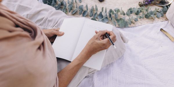 Person writing in a notebook while sitting outdoors on a blanket.