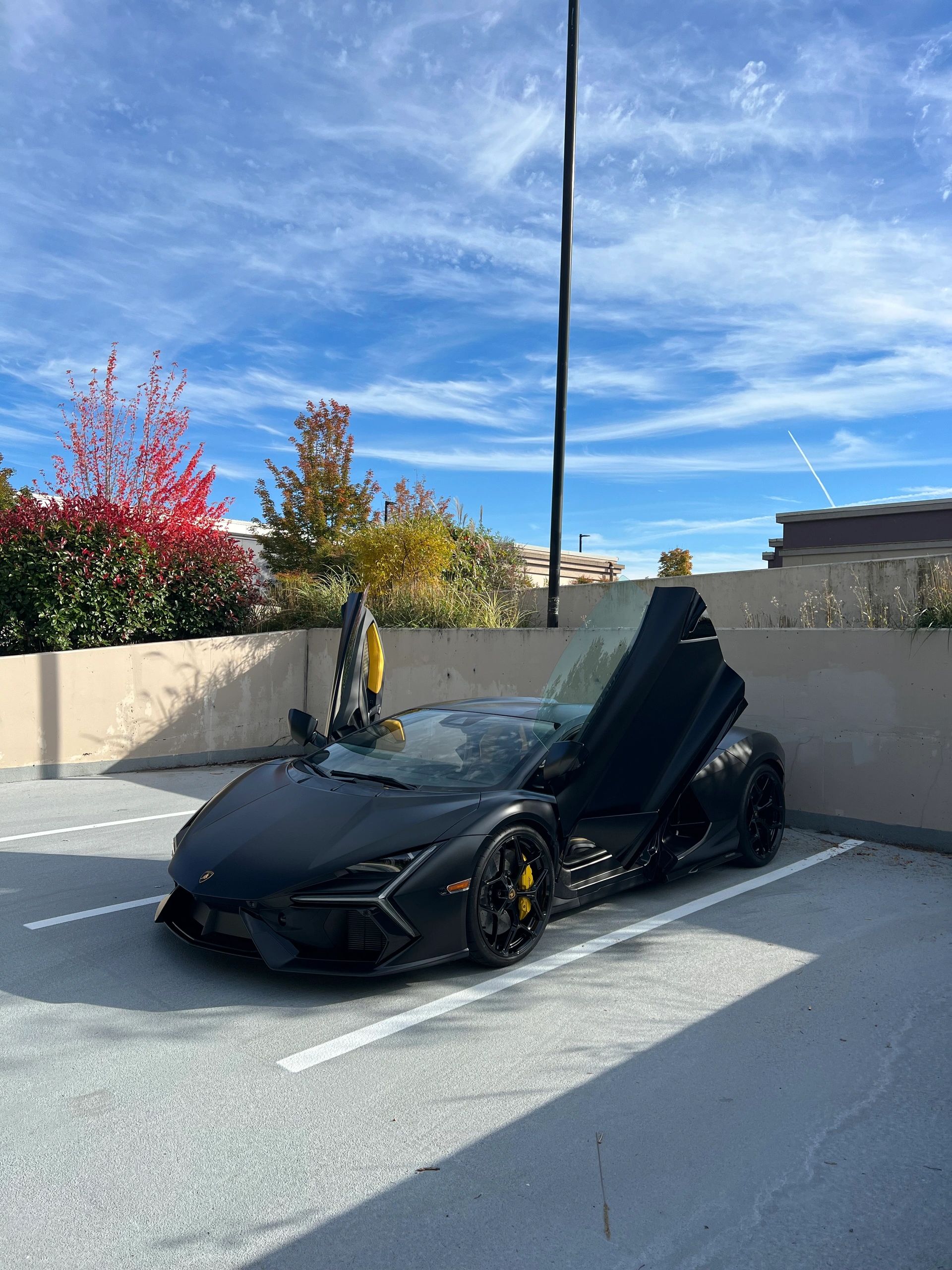 Black Lamborghini with scissor doors open in a parking lot under a blue sky.