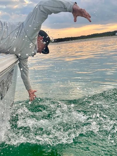 Man reaching over a boat into splashing green water at sunset.