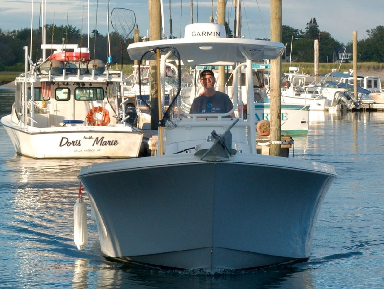 A man steering a boat in a calm marina with other boats docked nearby.