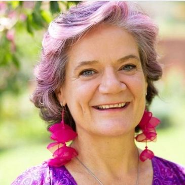 Smiling woman with pink-purple hair and vibrant earrings outdoors.