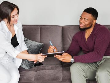 Man signing a document handed by a woman on a couch.